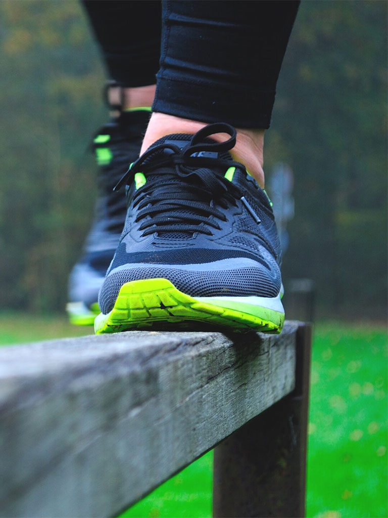 Person balanciert in Sportschuhen auf einem Holzgeländer im Freien, mit grünem Rasen und Bäumen im Hintergrund