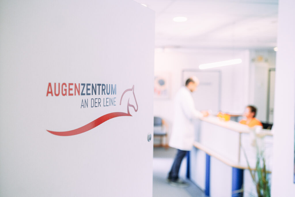 Eingangsbereich des Augenzentrums: geöffnete Tür mit Logo, dahinter Empfangssituation mit Arzt und Mitarbeiterin im Gespräch.